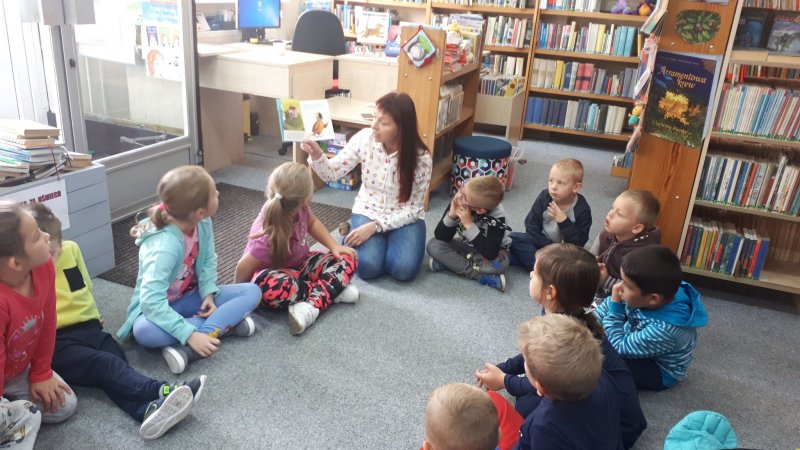 Zajęcia w bibliotece. Na podłodze siedzi grupa dzieci i młoda kobieta, która pokazuje dzieciom otwartą książkę. W głębi regały z książkami. Książki mają kolorowe grzbiety.