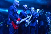 Na scenie w rzędzie stoją koncertujący muzycy z zespołu Poparzeni Kawą Trzy. Pierwszy z gitarą, kolejni z saksofonem i trąbką