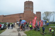 Pod Zamkiem Książąt Mazowieckich rowerzyści z rowerami, kilka samochodów, namiot i nadmuchiwana brama