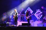 Zdjęcie obejmuje większy fragment sceny. Widać cały skład zespołu. Na pierwszym planie dwóch gitarzystów. Jeden z nich śpiewa