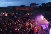Zdjęcie po zmroku. Scena na zamkowych błoniach. Trwa koncert. Przed sceną duża publiczność. W tle na wprost zamek Książąt Mazowieckich, po lewej w oddali parasole dla biesiadujących