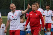 Piłkarze wchodzą na murawę. Każdy dorosły zawodnik idzie za rękę z małym piłkarzem. Jeden z zawodników z ręką wzniesioną do góry i uniesionym kciukiem