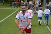 Przemysław Cypryański w piłkarskim stroju rozgrzewa się przed meczem. W oddali biegnie trzech małych zawodników piłki nożnej w sportowych strojach