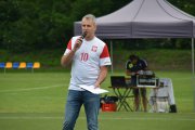 Komentator sportowy Jacek Kurowski mówi do mikrofonu. Za jego plecami widać rozstawiony na trawie namiot osłaniający urządzenia nagłaśniające