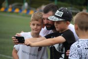 Dwóch małych chłopców ustawiło się do zdjęcia z Szymonem Kołeckim. Szymon pochylił się tak, żeby jego twarz znalazła się na wysokości twarzy chłopców. Jeden z tych chłopców robi zdjęcie telefonem