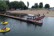 Zdjęcie wykonane z góry. Po lewej woda, ludzie pływają na rowerach wodnych. Pośrodku pomost, na którym stoi sporo osób. Po prawej piaszczysta plaża, po której chodzi kilka osób. W oddali parasole, pod którymi można posiedzieć