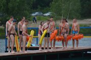 Na pomoście stoi grupa ratowników wodnych przygotowanych do pływania. W rękach trzymają koła i pasy ratownicze 