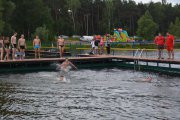 Trzy osoby skoczyły z nowego pomostu na kąpielisku Krubin. Kilka osób w strojach kąpielowych stoi na pomoście. W oddali, pod lasem widać nadmuchiwane zjeżdżalnie dla dzieci i kilka samochodów