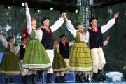 Występ zespołu ludowego. Na pierwszym planie dwie pary taneczne. Mężczyźni jedną ręką obejmują partnerki, drugą rękę unoszą w górę