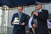 Na scenie Krzysztof Kosiński i Waldemar Pawlak. W. Pawlak mówi do mikrofonu. Ubrany jest w mundur strażacki. K. Kosiński, ubrany w garnitur, trzyma w rękach strażacki hełm