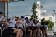 Na scenie gra orkiestra strażacka. W kadrze nastoletnie dziewczyny i chłopcy. W oddali widać miejski ratusz