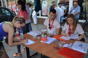 Punkt zapisów na bieg przeciwko białaczce. Za stołem siedzą wolontariusze zbierający zapisy. Z drupiej strony stołu, pochylona nad nim dziewczyna w sportowym stroju wypełnia kwestionariusz zapisu. Na stole stoi puszka na datki