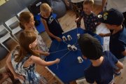 Wokół stolika stoją dzieci. Trzymają w rękach sznureczki, do których przyczepione są kubeczki stojące na stole. Kubeczki odwrócone są do góry dnem