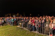 Za barierkami ochronnymi ustawionymi na trawie stoi wielu ludzi. To publiczność. W oddali po lewej przed barierkami dwie osoby siedzą na wózkach inwalidzkich