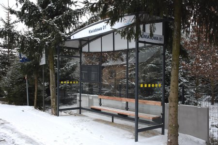 Wiata przystankowa przy ulicy pokrytej cienką warstwą śniegu. Po obu stronach nowej wiaty rosną bardzo wysokie iglaste drzewa. Wzdłuż krawędzi dachu wiaty znajduję się napis: Kwiatowa 1.