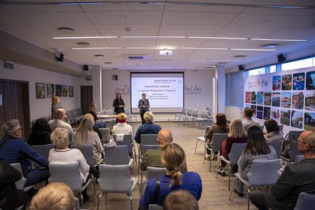 Spotkanie w ratuszu. Na sali wiele osob siedzących na krzesłach. W głębi ekran z wyświetloną informacją. W pobliżu ekranu stoją dwie kobiety.